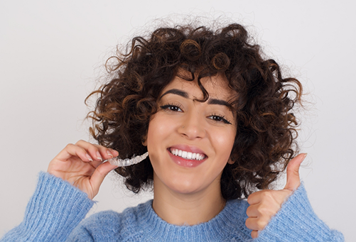 Woman in sweater holding clear aligner and giving thumbs up